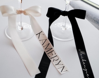 May include: Two champagne flutes with personalized ribbons tied around the stems. The white ribbon says "Kamerryn" and the black ribbon says "Mackenzie".