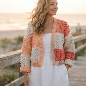 May include: A long-sleeve crochet cardigan featuring a color-blocked pattern of cream, peach, and coral yarn. The garment displays a textured design with alternating cable knit panels and raised bobble stitch details, finished with ribbed cuffs.