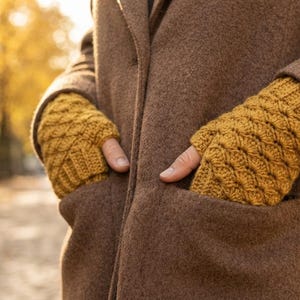 May include: A pair of mustard yellow fingerless gloves featuring a textured, raised shell stitch pattern and ribbed cuffs, worn with a brown wool coat. The gloves cover the hands while leaving the fingers exposed for dexterity and warmth.