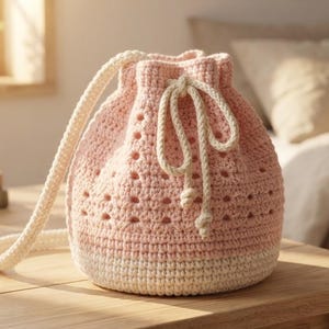 May include: A light pink and cream coloured crochet bucket bag featuring a textured stitch pattern with small openwork details. The bag includes a thick cream coloured drawstring closure and a matching long shoulder strap made of braided cord.