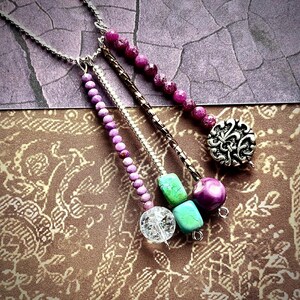 May include: Close-up of three necklaces with various beads and pendants. One necklace features a silver pendant with a floral design. Other necklaces have purple, green, and clear beads. The necklaces are on a patterned brown and purple background.