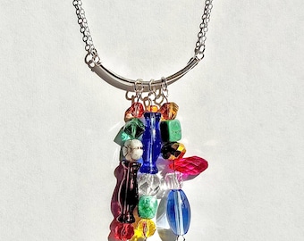 May include: A silver chain necklace with a curved bar pendant. The pendant features a cluster of colorful glass beads in various shapes and sizes, including red, blue, green, and yellow. The beads are strung together, creating a unique and eye-catching design.