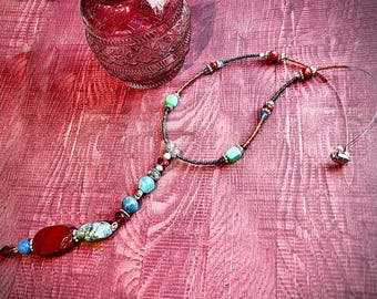 May include: A beaded necklace with a pendant featuring red, blue, and turquoise beads. The necklace has a silver chain and a small heart-shaped pendant. A decorative glass jar is in the background.