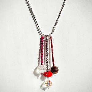 May include: A silver chain necklace with multiple strands of beads and crystal pendants. The strands include ruby-colored beads, silver beads, and dark red beads. The pendants are clear and red, with a faceted crystal at the bottom.