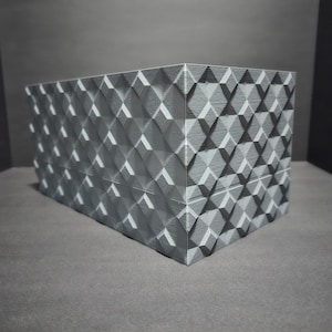 May include: A gray box with a geometric pattern on all sides. The box is made of a textured material and has a slightly raised design.