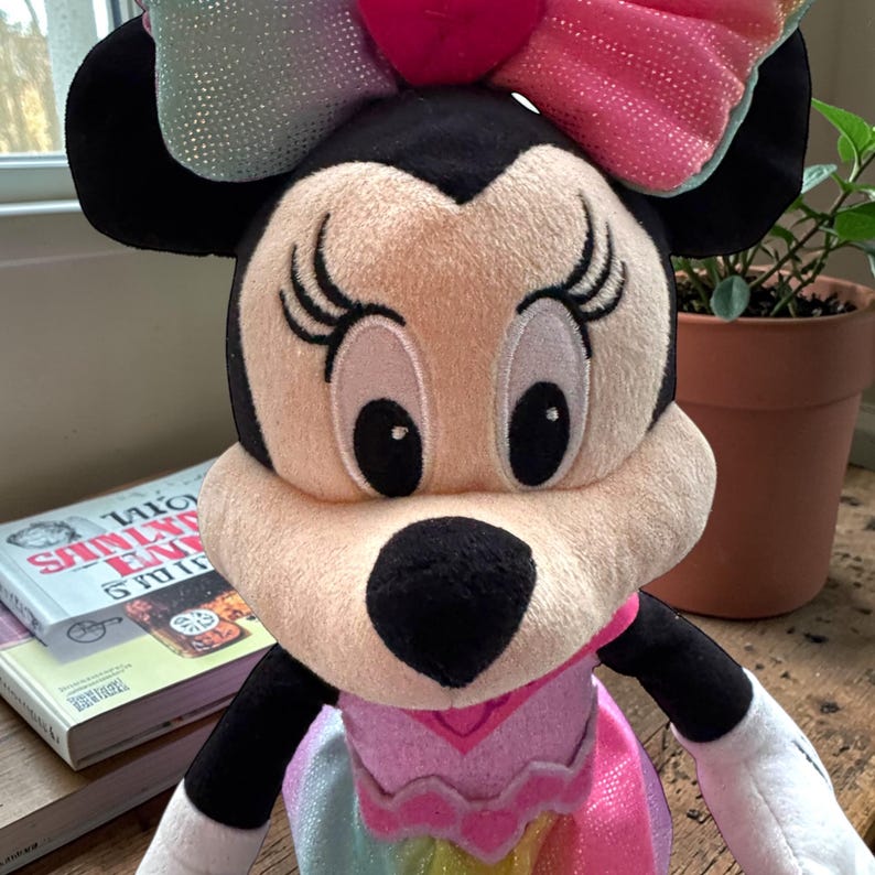 A plush toy of Minnie Mouse, with a bow on her head, is sitting on a wooden surface next to a potted plant.