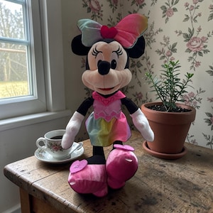 A Minnie Mouse plush toy is sitting on a wooden table next to a potted plant and a cup of coffee.