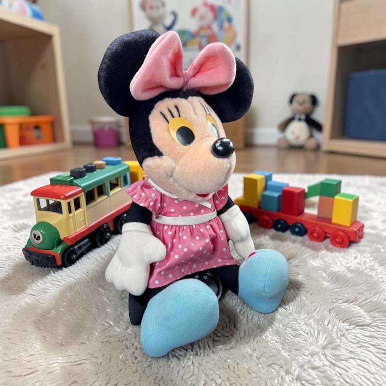 A Minnie Mouse plush toy is sitting on a carpet, surrounded by colorful building blocks and a toy train.