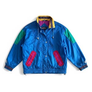 May include: A blue windbreaker jacket featuring a yellow collar with a patterned trim, green shoulder panels, and two vertical pink zippered pockets. The jacket includes a front snap button closure, a purple inner lining, and patterned cuffs.