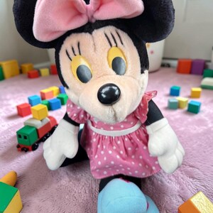 A stuffed Minnie Mouse toy wearing a pink polka dot dress sits on a pink blanket surrounded by colorful building blocks.