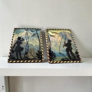 May include: Two framed silhouette art pieces. One depicts a person with a bucket, the other a person fishing. Both have a gold and brown striped border and a colorful mountain backdrop.