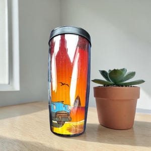 A colorful glass mug with a picture of a car and a bird on it, placed next to a small potted succulent plant on a wooden surface.