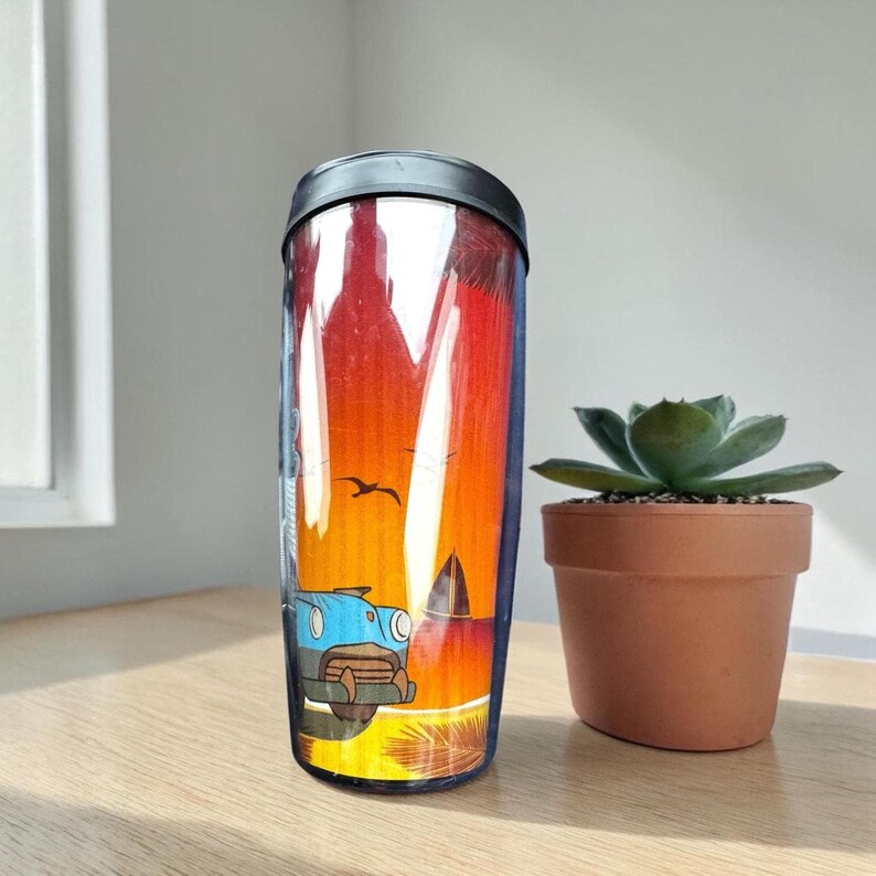 A colorful glass mug with a picture of a car and a bird on it, placed next to a small potted succulent plant on a wooden surface.