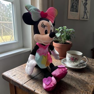 A Minnie Mouse plush toy is sitting on a wooden table next to a cup of tea and a potted plant.