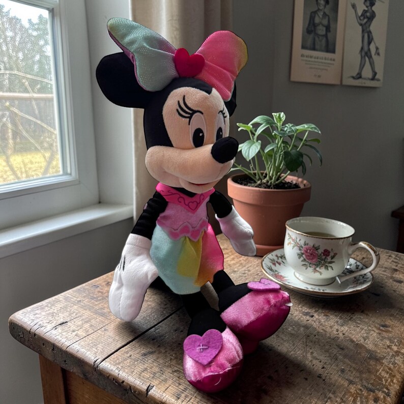A Minnie Mouse plush toy is sitting on a wooden table next to a cup of tea and a potted plant.