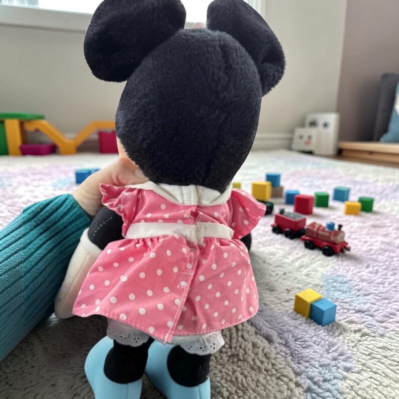 A plush toy of Minnie Mouse dressed in a pink polka dot dress is sitting on a carpet, surrounded by colorful blocks and toys.