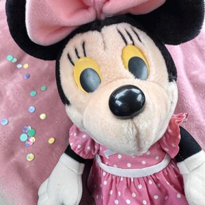 A close-up of a Minnie Mouse plush toy, with its large yellow eyes and black nose, wearing a pink dress with white polka dots. The toy is placed on a pink surface with scattered colorful buttons around it.