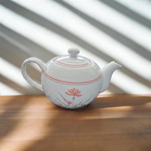 May include: A white ceramic teapot featuring a rounded body, a matching lid with a small knob, and a curved handle. The teapot is decorated with a delicate pink floral pattern and thin pink horizontal bands encircling the upper portion.
