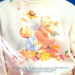 a person wearing a white sweatshirt with a colorful illustration of two angels, one in a pink dress and the other in a blue dress, surrounded by autumn leaves.