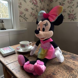 A plush toy of Minnie Mouse, dressed in a colorful outfit, sits on a wooden table next to a teacup and a book.