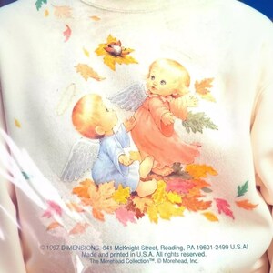 a person wearing a white sweatshirt with a colorful illustration of two angels, one in a pink dress and the other in a blue dress, surrounded by autumn leaves.