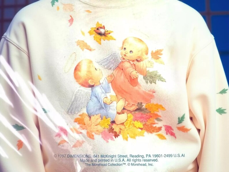 a person wearing a white sweatshirt with a colorful illustration of two angels, one in a pink dress and the other in a blue dress, surrounded by autumn leaves.