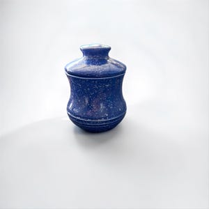 Peut inclure: Un pot en céramique bleu cobalt avec un couvercle assorti, présentant un motif blanc moucheté sur toute la surface. Le pot a une silhouette incurvée en forme de sablier avec de fines stries horizontales près de la base et un bouton arrondi.