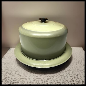May include: A vintage, two-toned cake carrier in shades of green and cream. The carrier has a domed lid with a black knob and sits on a matching circular base. The design is simple and functional, likely from the mid-20th century.