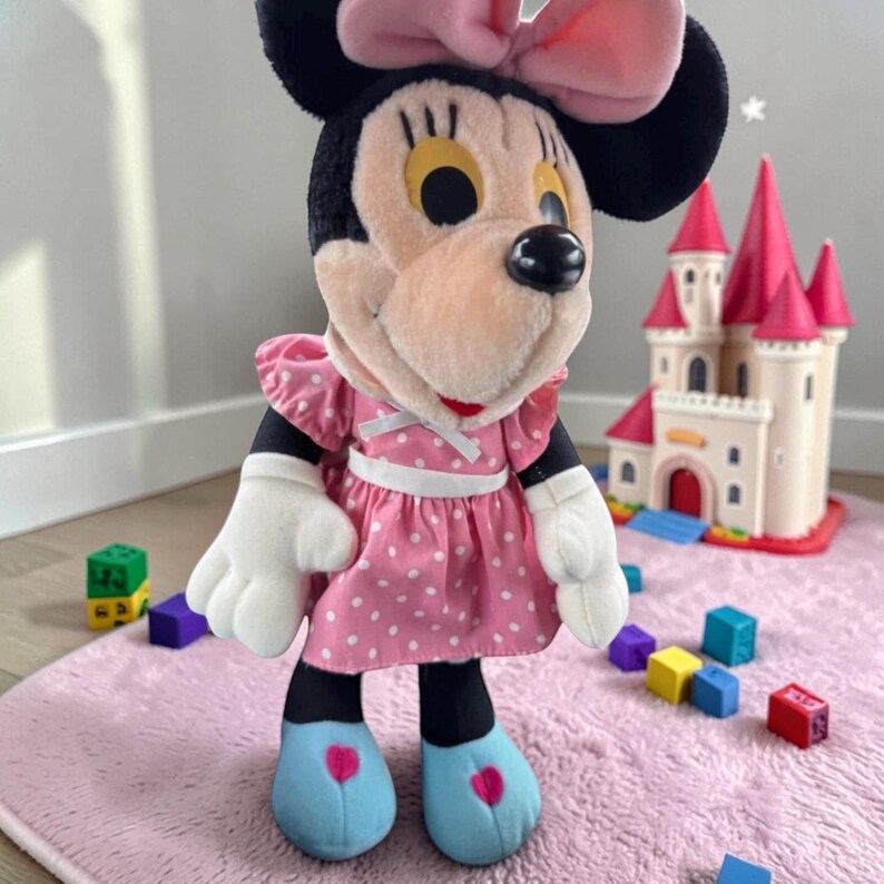A Minnie Mouse plush toy wearing a pink dress and blue shoes, standing on a pink rug with colorful blocks in the background.