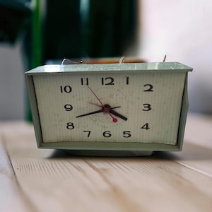 May include: A rectangular light green analog alarm clock with a textured off-white face. The clock features black Arabic numerals from one to twelve, black hour and minute hands, and a thin red second hand with a small red circular accent.