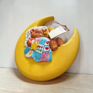 May include: A yellow crescent moon wall plaque featuring a brown teddy bear tucked under a colorful patchwork quilt with red hearts and tulips. The bear holds a yellow star, and a white rectangular tag is attached by a thin brown string.