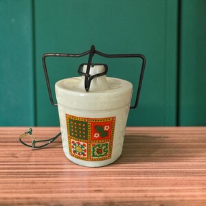 May include: A cream-colored ceramic jar with a black metal wire handle and lid locking mechanism. The front features a square grid pattern in red, green, and yellow, containing small floral and geometric motifs. A thin green wire is attached to the side.