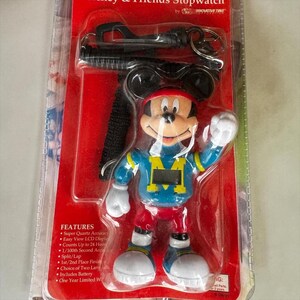 May include: Mickey & Friends stopwatch in packaging. The stopwatch is shaped like Mickey Mouse, wearing a blue and yellow jersey and red trousers. The packaging is red with black text and features. Includes a black lanyard and keychain.