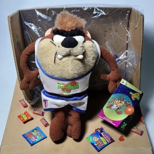 May include: A plush Taz character from Space Jam, wearing a white and blue basketball uniform. The plush toy is brown, tan, and black, with a tag featuring Taz. Candy wrappers are scattered around the toy.
