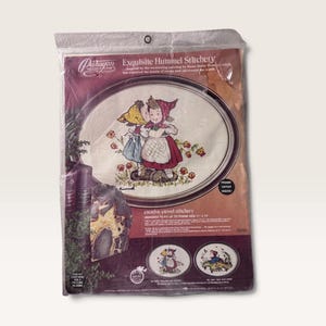 May include: A Paragon Needlecraft Exquisite Hummel Stitchery crewel kit in a clear plastic package. The packaging features an oval design of two children, one in a red bonnet and one in a yellow bonnet, with the text "Designed to fit up to frame size 11" x 14"."