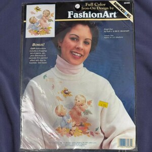 The image is a package for Iron-On Design by FashionArt, featuring a smiling woman with a white sweater adorned with a design of cherubs and leaves.