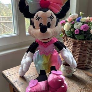 A Minnie Mouse plush toy is sitting on a wooden table next to a basket filled with colorful flowers.