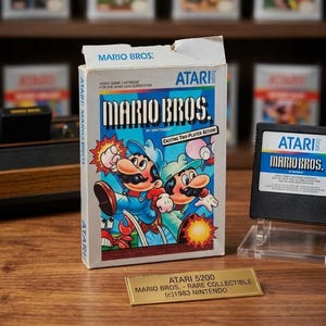 May include: A white cardboard box for the Atari 5200 Mario Bros. video game cartridge featuring illustrations of Mario and Luigi. Next to it is the black game cartridge and a gold plaque reading ATARI 5200 MARIO BROS. - RARE COLLECTIBLE (c)1983 NINTENDO.