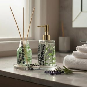 May include: A clear glass soap dispenser with a gold pump and a matching reed diffuser set on a marble countertop. Both are decorated with lavender and green leaf designs. A stack of white towels is in the background.