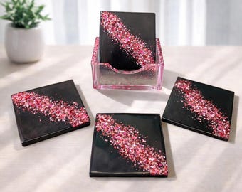 Puede incluir: Juego de cuatro posavasos cuadrados negros con una franja diagonal de purpurina rosa. Los posavasos se muestran con un soporte a juego. Una pequeña planta en maceta está en el fondo.