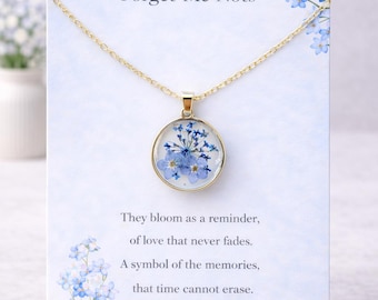May include: A gold-colored necklace with a round pendant featuring blue forget-me-not flowers encased in clear resin. The pendant hangs on a delicate chain. The card behind the necklace reads "Forget-Me-Nots".