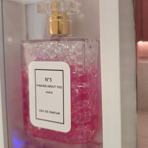 K&ouml;nnte beinhalten: Ein rechteckiger Parf&uuml;mflakon mit einem transparenten, strukturierten K&ouml;rper und einem goldfarbenen Verschluss. Die Flasche ist mit rosa Fl&uuml;ssigkeit gef&uuml;llt und hat ein wei&szlig;es Etikett mit dem Text "N&deg;5 PINKING ABOUT YOU PARIS EAU DE PARFUM". Die Flasche wird in einer wei&szlig;en Box pr&auml;sentiert.