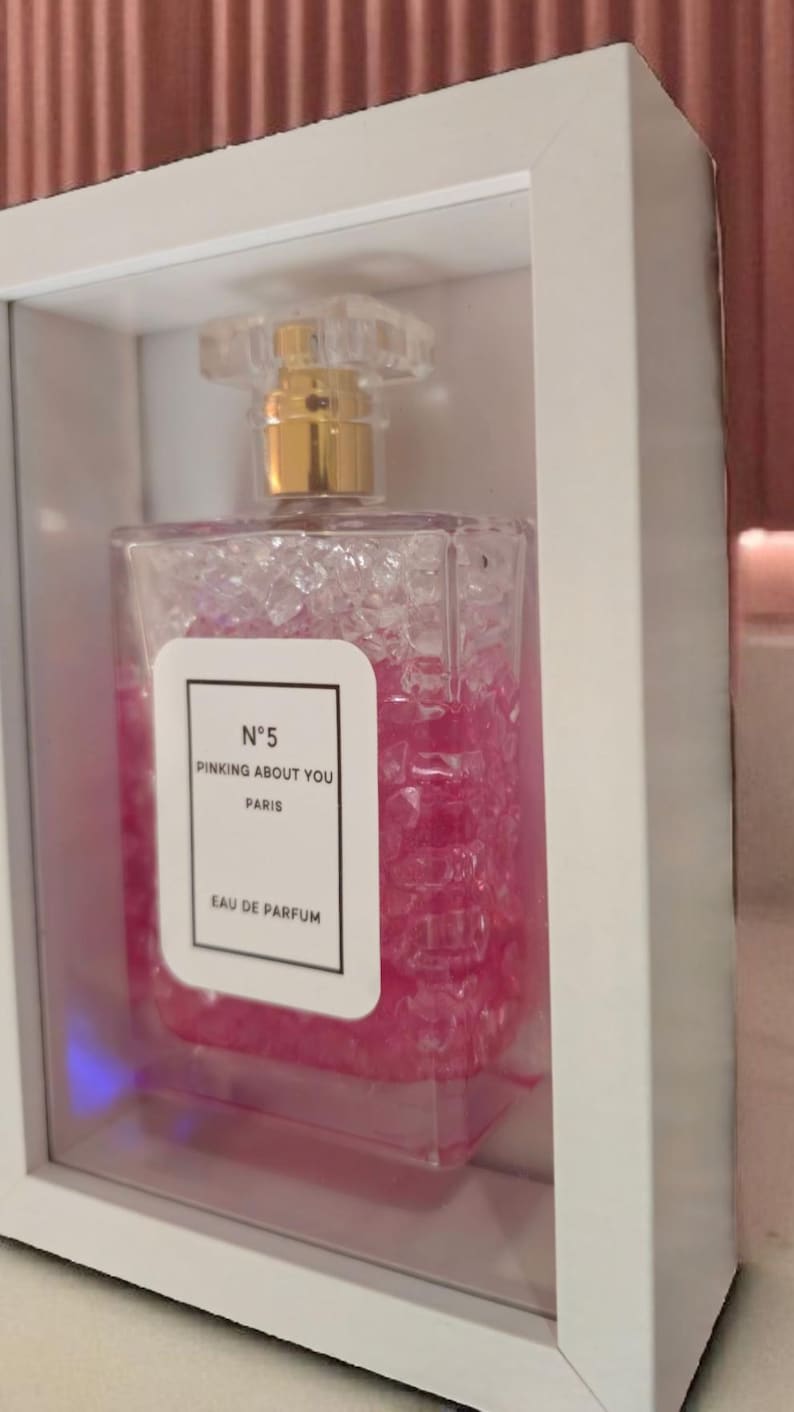 K&ouml;nnte beinhalten: Ein rechteckiger Parf&uuml;mflakon mit einem transparenten, strukturierten K&ouml;rper und einem goldfarbenen Verschluss. Die Flasche ist mit rosa Fl&uuml;ssigkeit gef&uuml;llt und hat ein wei&szlig;es Etikett mit dem Text "N&deg;5 PINKING ABOUT YOU PARIS EAU DE PARFUM". Die Flasche wird in einer wei&szlig;en Box pr&auml;sentiert.