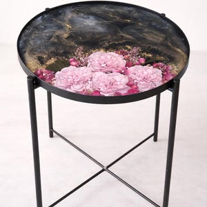 Puede incluir: Mesa auxiliar redonda de metal negro. La tapa presenta un diseño veteado negro y dorado con flores rosas. La mesa tiene un diámetro aproximado de 45 cm.