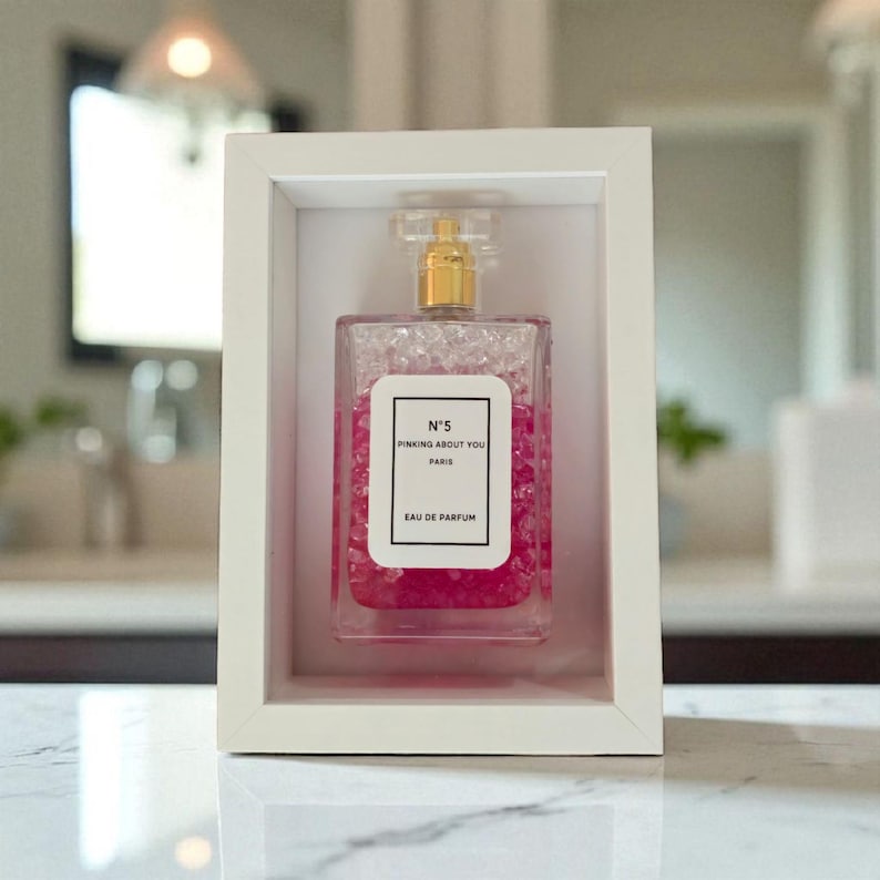 K&ouml;nnte beinhalten: Ein gerahmter Parf&uuml;mflakon mit goldenem Verschluss und dem Etikett "N&deg;5 Pinking About You Paris Eau De Parfum". Der Flakon ist mit rosa Fl&uuml;ssigkeit und transparenten Perlen gef&uuml;llt.