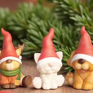Peut inclure: Trois figurines de Noël : un renne, un chat et un chien, chacun portant un bonnet de Père Noël rouge et blanc. Le renne a des bois et une écharpe verte. Le chat et le chien sont de couleur claire. Les figurines sont sur fond de branches de pin.