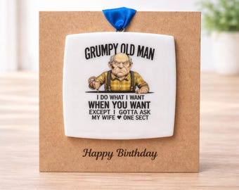 Op de afbeelding: Een wit keramisch ornament met de tekst "GRUMPY OLD MAN" en een cartoon illustratie van een chagrijnige man. Het ornament bevat ook de tekst "I DO WHAT I WANT WHEN YOU WANT EXCEPT I GOTTA ASK MY WIFE ONE SECT." Op een bruine kaart met "Happy Birthday".