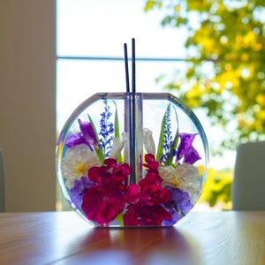 Op de afbeelding: Een heldere, ronde harsvaas met een verscheidenheid aan kleurrijke bloemen erin. De bloemen omvatten paarse irissen, witte en paarse viooltjes en rode bloemen. Twee zwarte stokjes zijn in het midden van de vaas geplaatst.