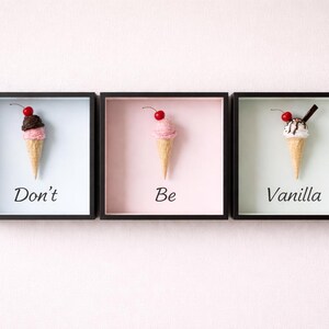May include: Three framed art pieces with ice cream cone sculptures. Each cone has a different topping and a cherry. The text reads "Don't Be Vanilla". The frames are black with different pastel backgrounds.