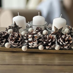 Può includere: Un centrotavola decorativo con tre candele bianche in portacandele di vetro, circondato da pigne, sfere bianche e ornamenti a forma di stella. La composizione è su una base di legno, ideale per le festività.